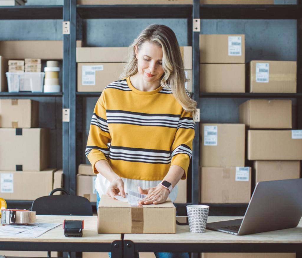 Woman in a striped sweater packing a cardboard parcel at a desk beside shelves of boxes and a laptop in a fulfillment workspace