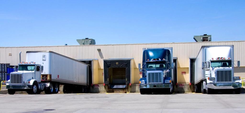 Three semi-trucks backed into loading docks of a tan warehouse under clear blue sky