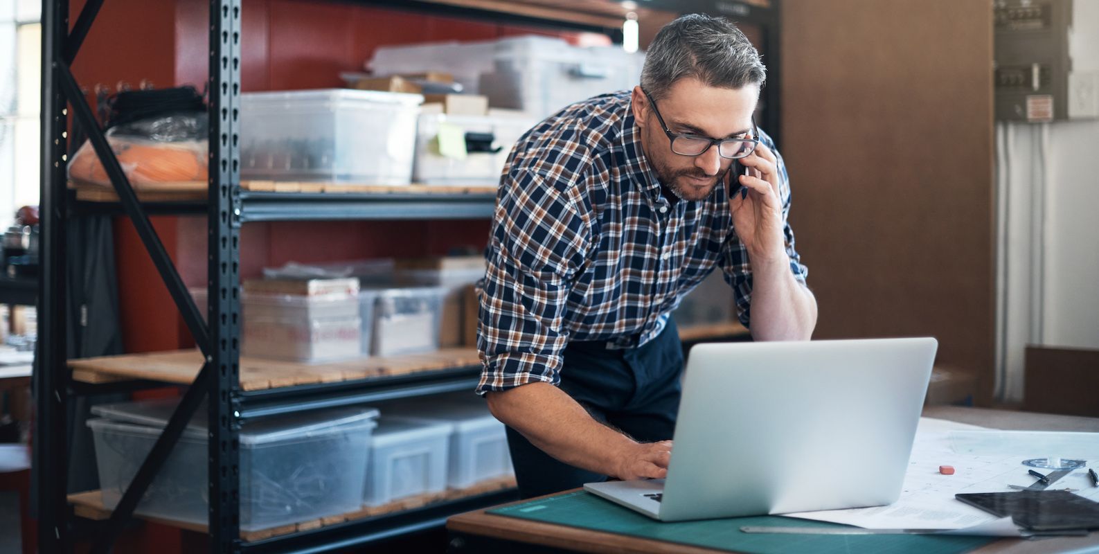 Man in plaid shirt and glasses talks on a phone while using a laptop beside storage shelves