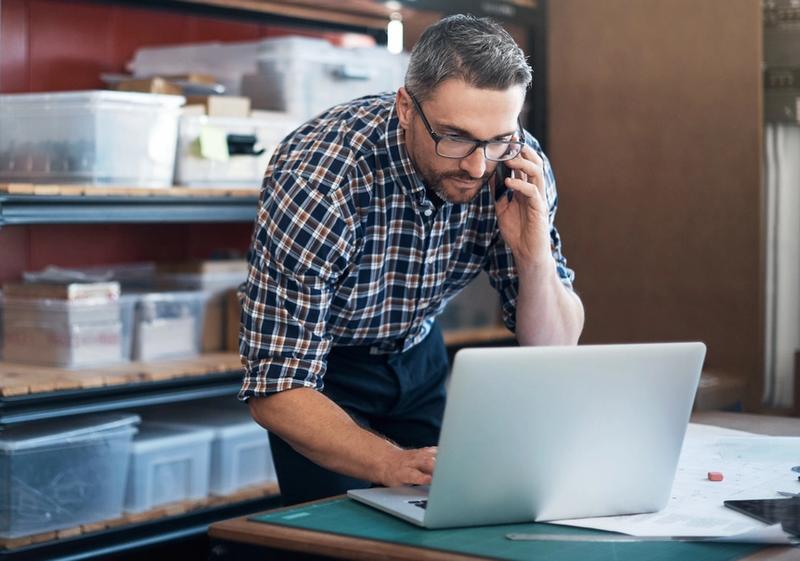 Man in plaid shirt and glasses talks on a phone while using a laptop beside storage shelves