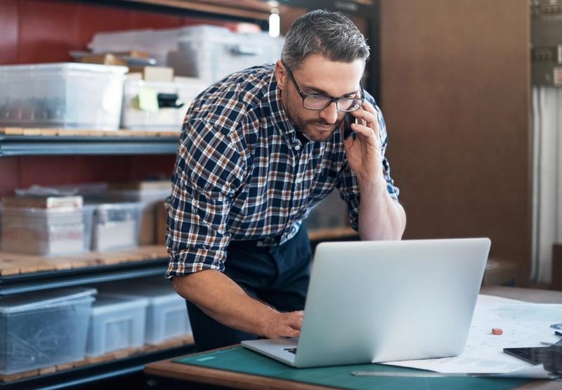 Man in plaid shirt and glasses talks on a phone while using a laptop beside storage shelves