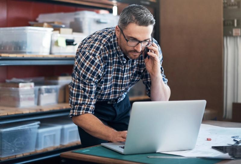 Man in plaid shirt and glasses talks on a phone while using a laptop beside storage shelves