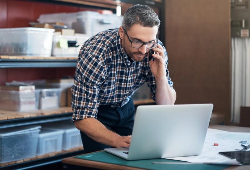 Man in plaid shirt and glasses talks on a phone while using a laptop beside storage shelves