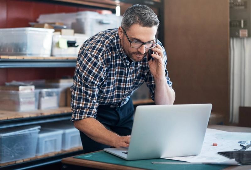 Man in plaid shirt and glasses talks on a phone while using a laptop beside storage shelves