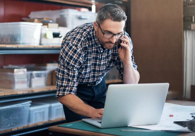 Man in plaid shirt and glasses talks on a phone while using a laptop beside storage shelves