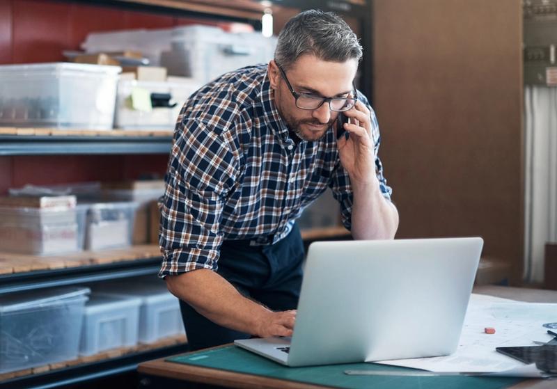 Man in plaid shirt and glasses talks on a phone while using a laptop beside storage shelves