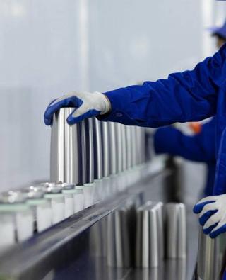 Worker in blue uniform and gloves inspecting stainless steel cups on a factory production line