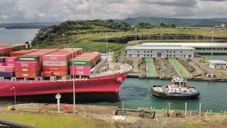A container ship loaded with stacked containers sits in a Panama Canal lock chamber as a tugboat waits alongside in the green water.