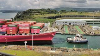 A container ship loaded with stacked containers sits in a Panama Canal lock chamber as a tugboat waits alongside in the green water.