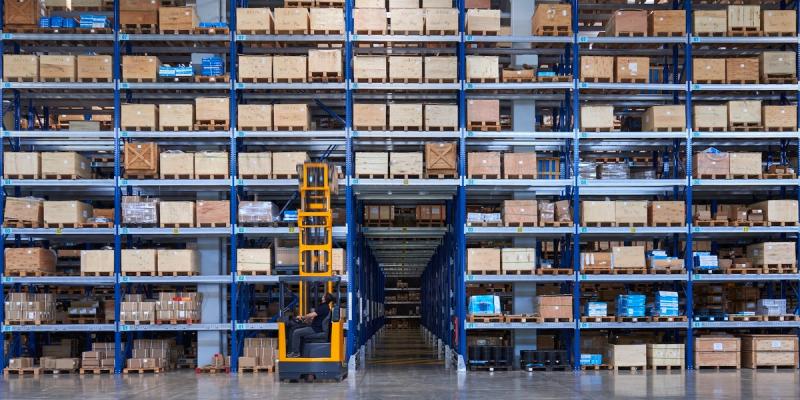 High-bay warehouse with tall blue racks of pallets and a worker on an order-picker lift in the aisle