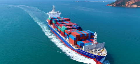 Aerial photo of a fully loaded container ship underway on open water, leaving a long wake, with a rocky headland in the distance.