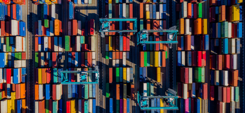 Overhead aerial photo of tightly packed rows of multicolored shipping containers with teal gantry cranes moving between them.