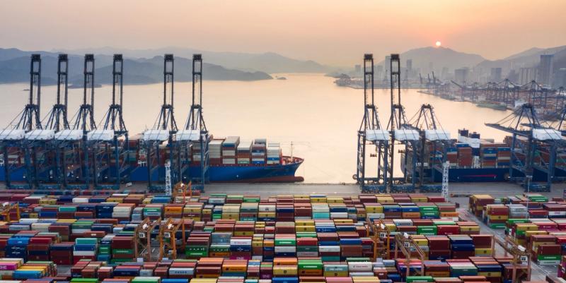 Aerial view of a container port at sunset with gantry cranes, docked cargo ships, and rows of stacked containers