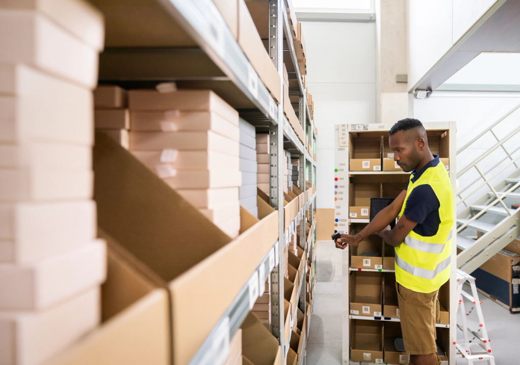 Warehouse worker in safety vest checking items on shelves while holding a handheld device.