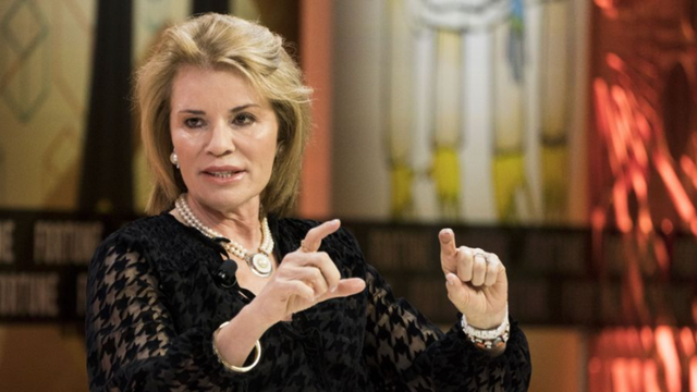 Woman with blond hair in a black lace top gesturing with both hands during an on-stage interview