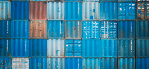 Photo of a wall of stacked blue and weathered shipping containers filling the frame at a container yard.