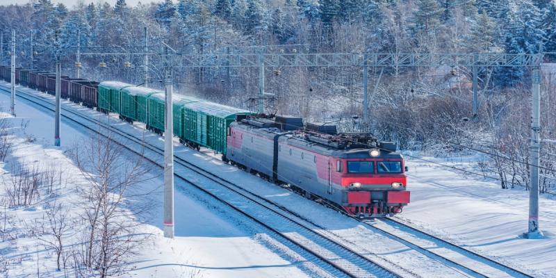 Red and gray electric locomotive hauls green and dark freight cars on snowy parallel tracks in winter