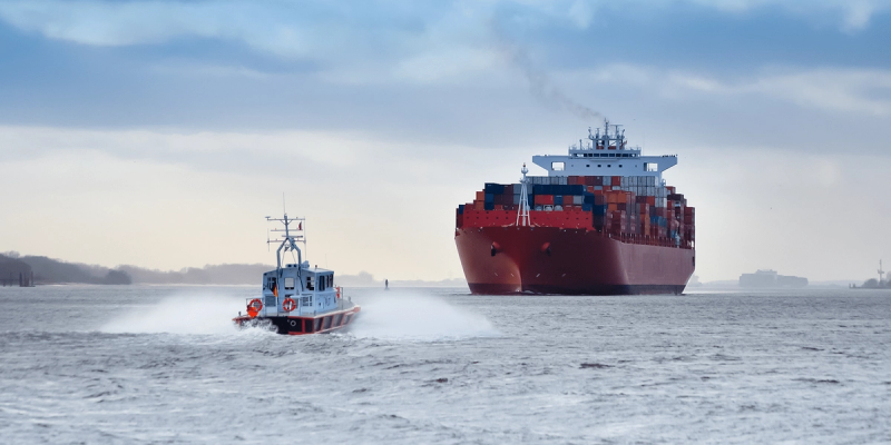 Red container ship escorted by a pilot boat across choppy gray water under an overcast sky