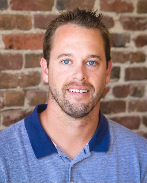 Headshot of a smiling man with short hair in a blue polo shirt against an exposed brick wall