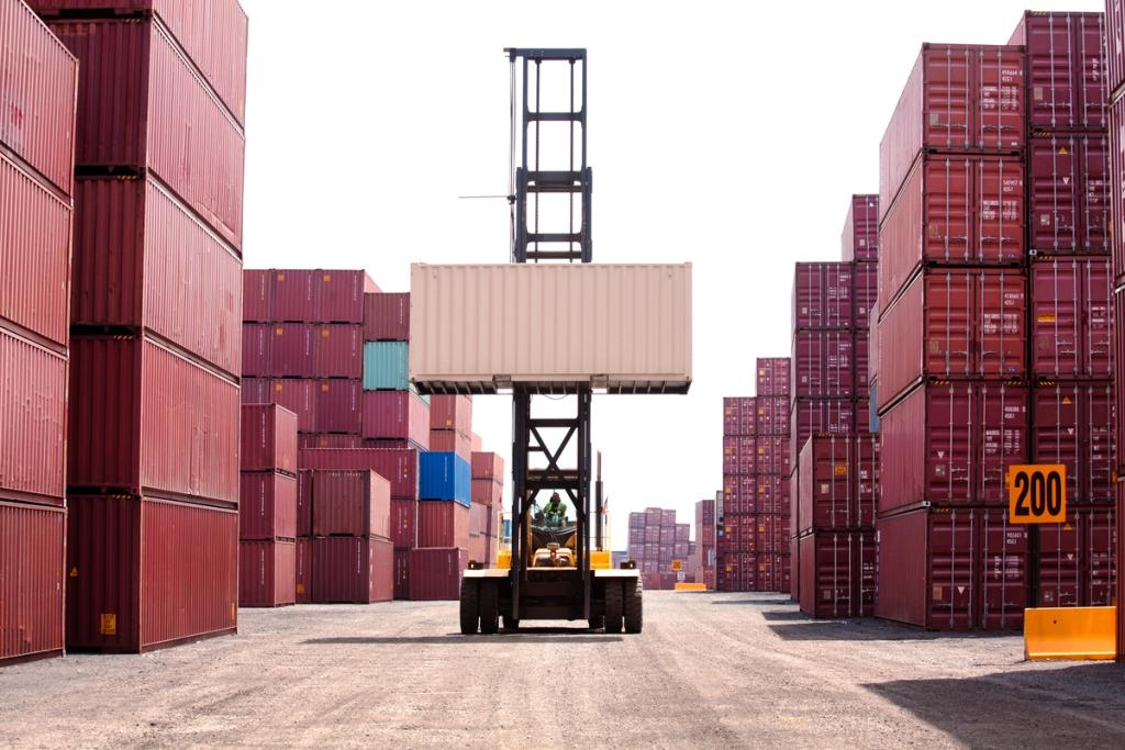 Reach stacker lifting a shipping container high in a container yard lined with stacks of maroon containers under a bright sky