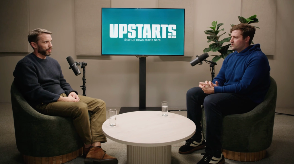 Two men seated in chairs during a filmed interview, with a screen displaying the Upstarts Media logo between them