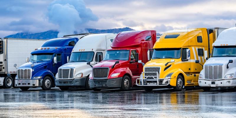 Row of parked semi truck cabs in blue, white, red, yellow and white on a wet lot with mountains behind