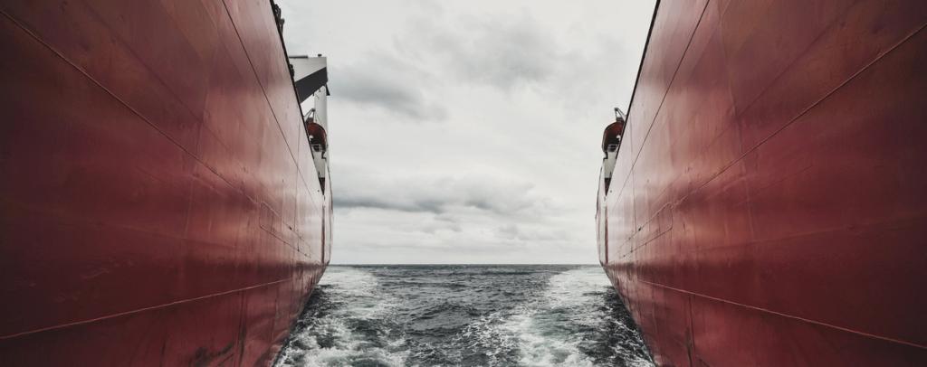 View between the towering red hulls of two ships, churning sea and grey horizon visible in the gap.