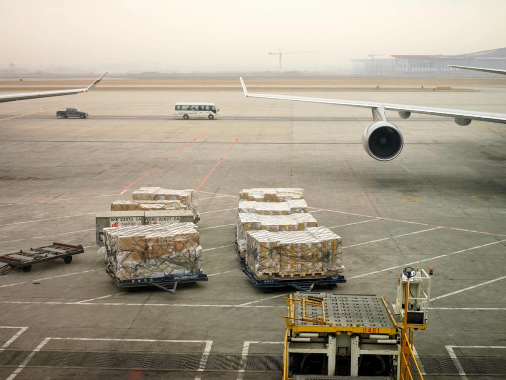 Netted air cargo pallets on the apron beside an aircraft wing and engine at a hazy airport