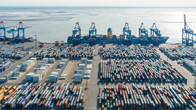 Aerial view of a container port with rows of stacked shipping containers, gantry cranes, and a docked cargo ship beside the water
