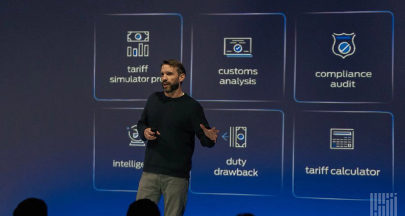 Speaker on stage gesturing in front of a screen showing tariff simulator, customs analysis, compliance audit and duty drawback panels