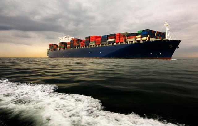 Container ship fully loaded with stacked containers sailing under a cloudy sky, wake in the foreground