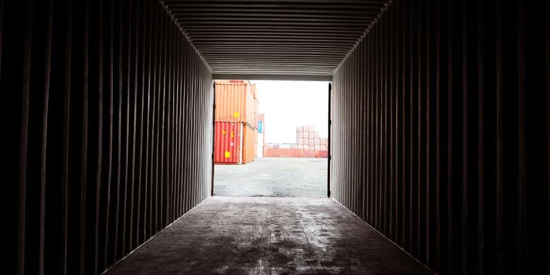 View out of a dark shipping container toward orange and red containers stacked in a port yard.