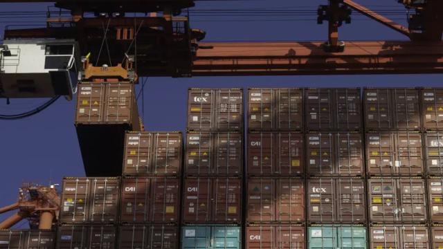 Gantry crane lifting a shipping container above stacked rows of cargo containers at a port under a blue sky