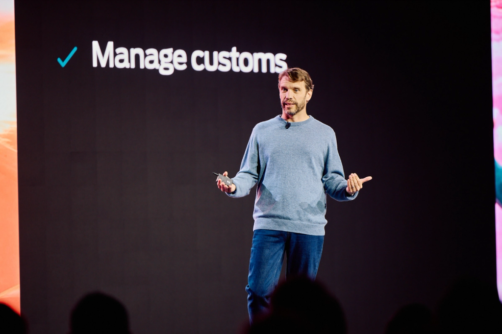 Man in a blue sweater speaks on stage in front of a screen reading Manage customs with a checkmark