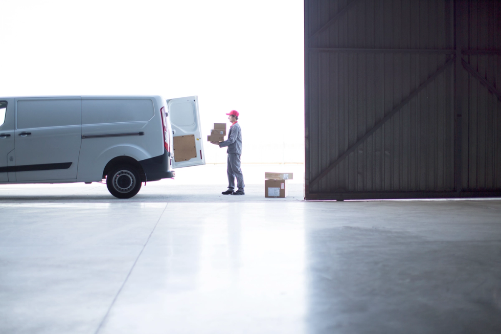 Courier in red cap loads parcels from an open white delivery van into a dark warehouse doorway.