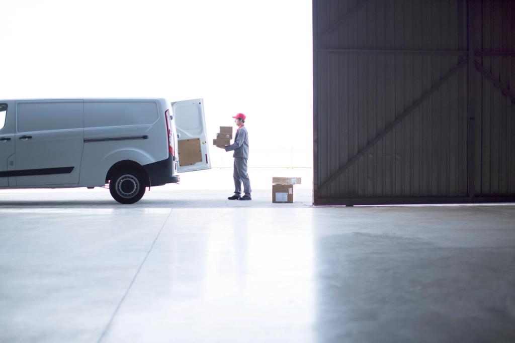 Courier in red cap loads parcels from an open white delivery van into a dark warehouse doorway.