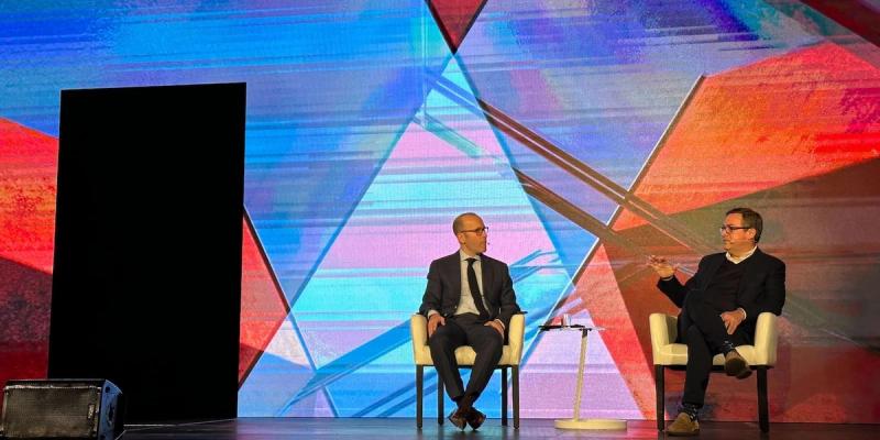 Two men in suits seated on stage for a fireside chat against a colorful geometric LED backdrop