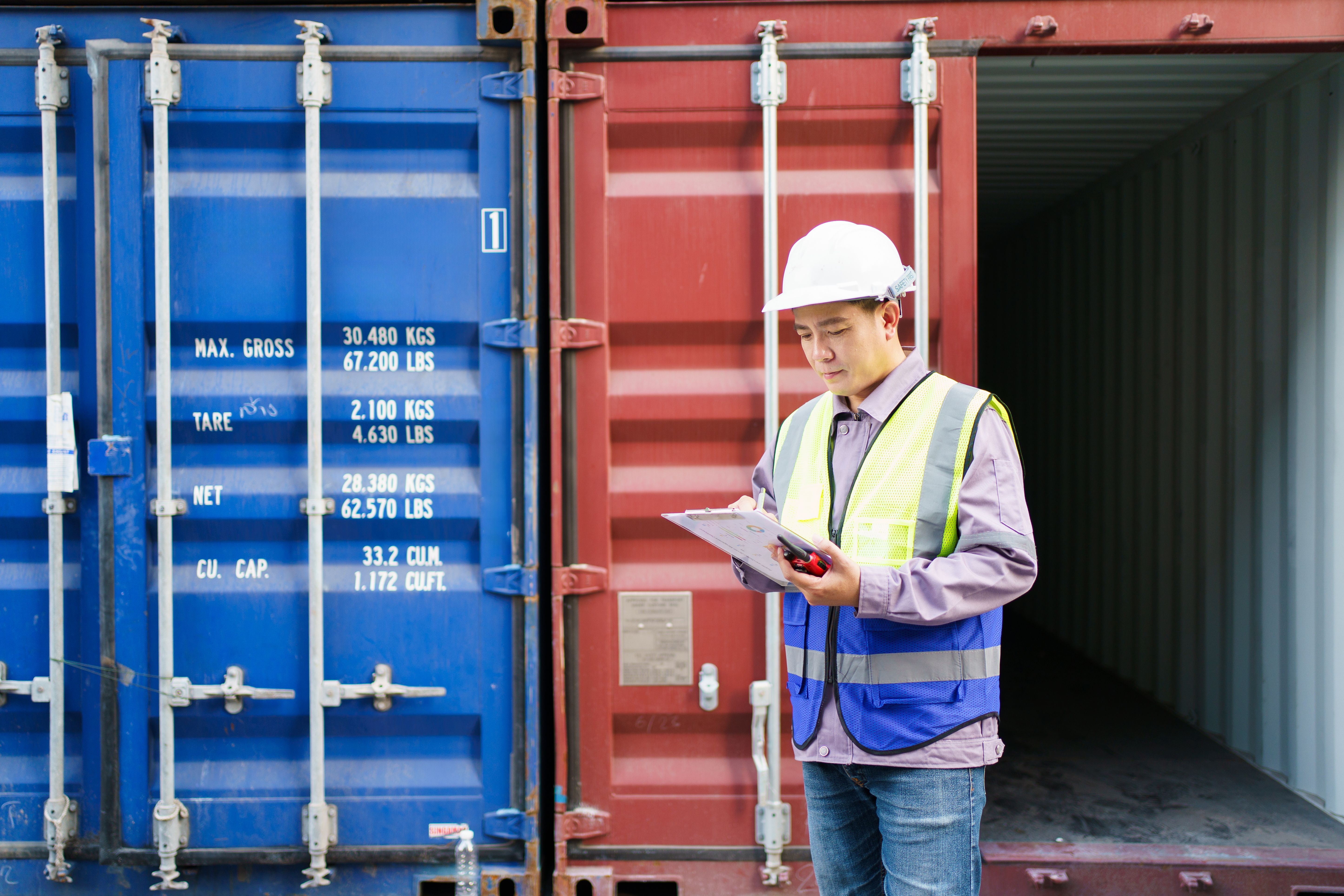 Worker in hard hat and safety vest inspecting shipping containers with a clipboard