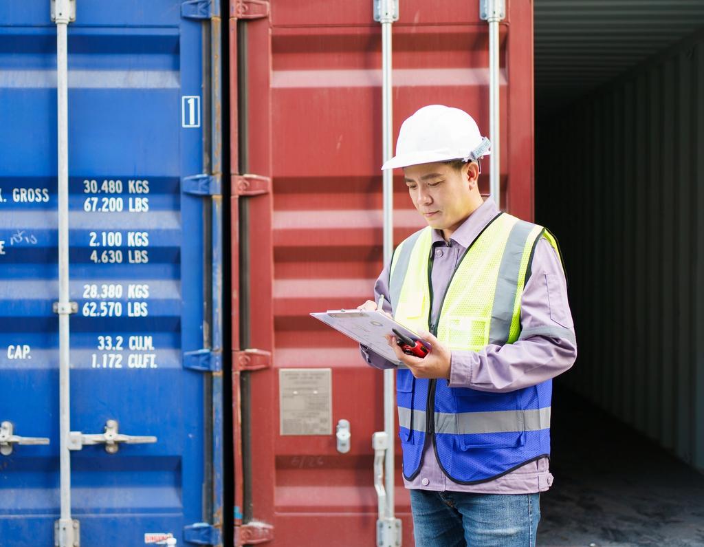 Worker in hard hat and safety vest inspecting shipping containers with a clipboard