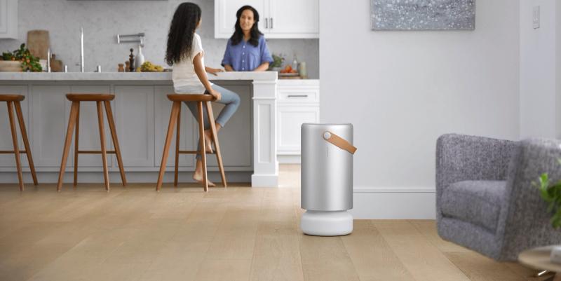 Silver Molekule air purifier on a wood floor with two women chatting at a kitchen island behind
