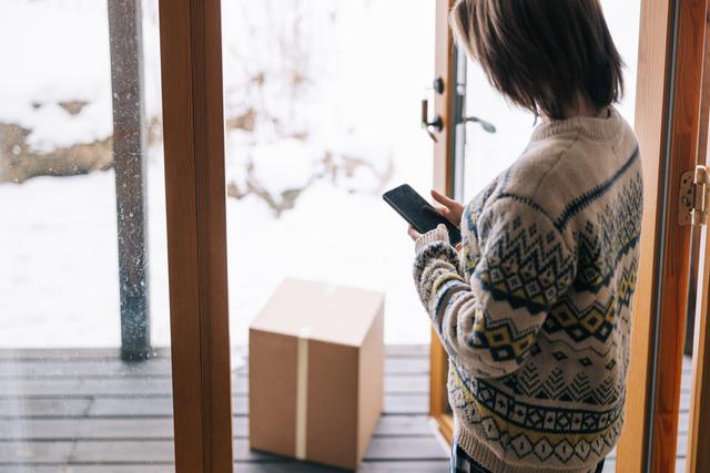 Person in patterned sweater checks a phone at an open door with a parcel on a snowy porch