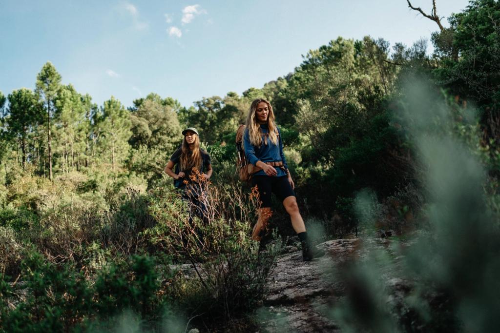 Two women in outdoor apparel stand on a rocky trail surrounded by green brush and trees under a blue sky