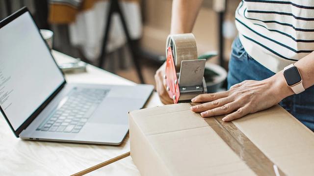 Person sealing a cardboard shipping box with packing tape at a desk beside a laptop