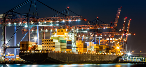 Night photo of a container ship being worked under floodlit cranes at a brightly lit port terminal.