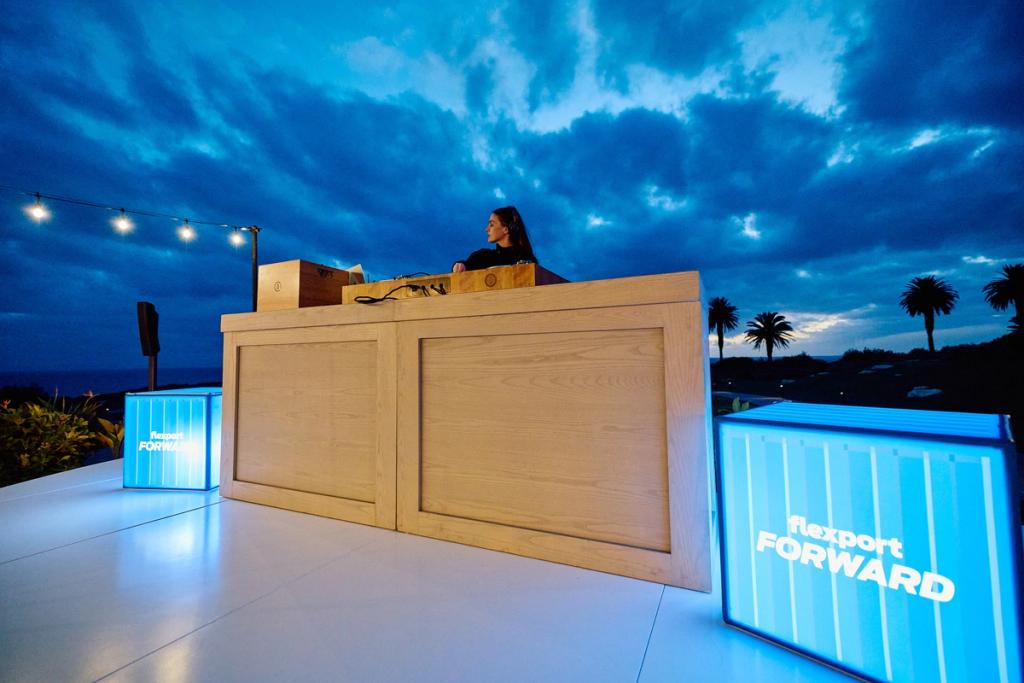 Person at a wooden DJ booth at dusk beside glowing Flexport FORWARD light boxes on an outdoor patio