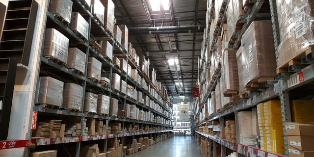 Long aisle of a high-bay warehouse with tall racks of palletized boxes, evoking inventory and ERP-driven logistics.