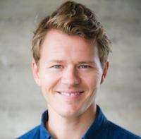 Smiling young man with short blond hair in a blue denim shirt, headshot against a concrete wall