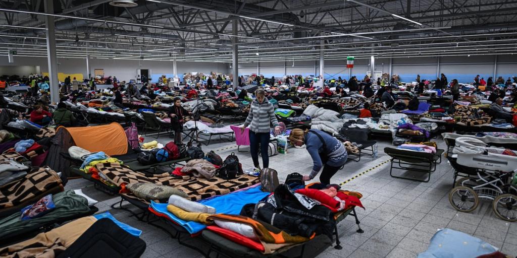 Crowded indoor refugee shelter filled with rows of cots, blankets and people