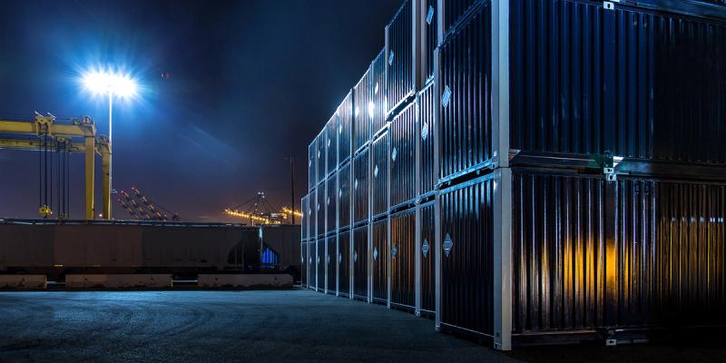 Stacks of shipping containers at a port terminal at night, lit by a floodlight with a crane in the distance