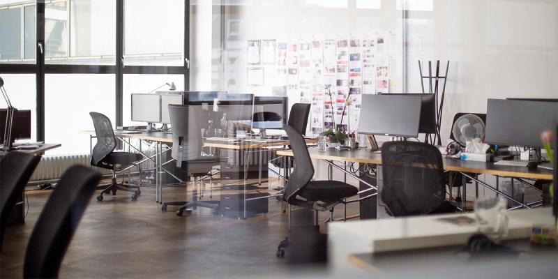 Empty modern open-plan office with rows of desks, monitors, mesh chairs and a wall of pinned papers
